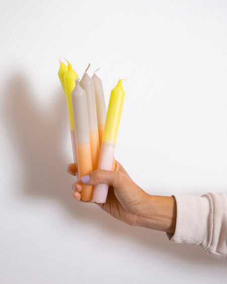 Hand hält vier bunte Kerzen, die aus Gelb, Grau und Rosa bestehen, vor einem weißen Hintergrund.
