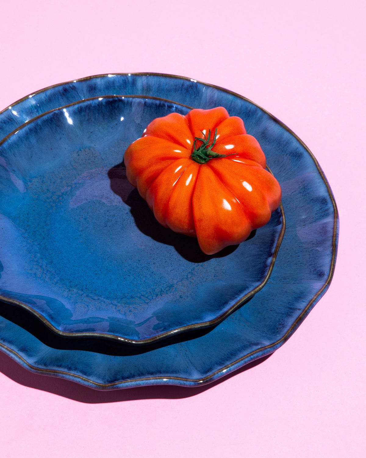 Zwei blaue Teller mit einer großen, gerippten Tomate darauf, auf einem rosa Hintergrund.