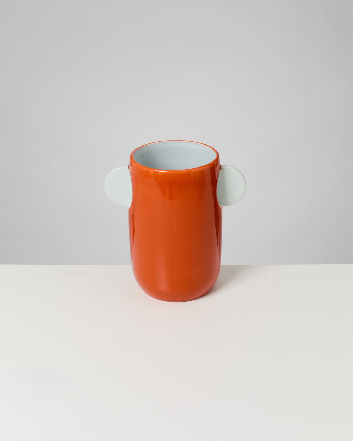 Eine orange Vase mit zwei seitlichen, weißen Griffen steht vor einem grauen Hintergrund auf einem weißen Tisch.