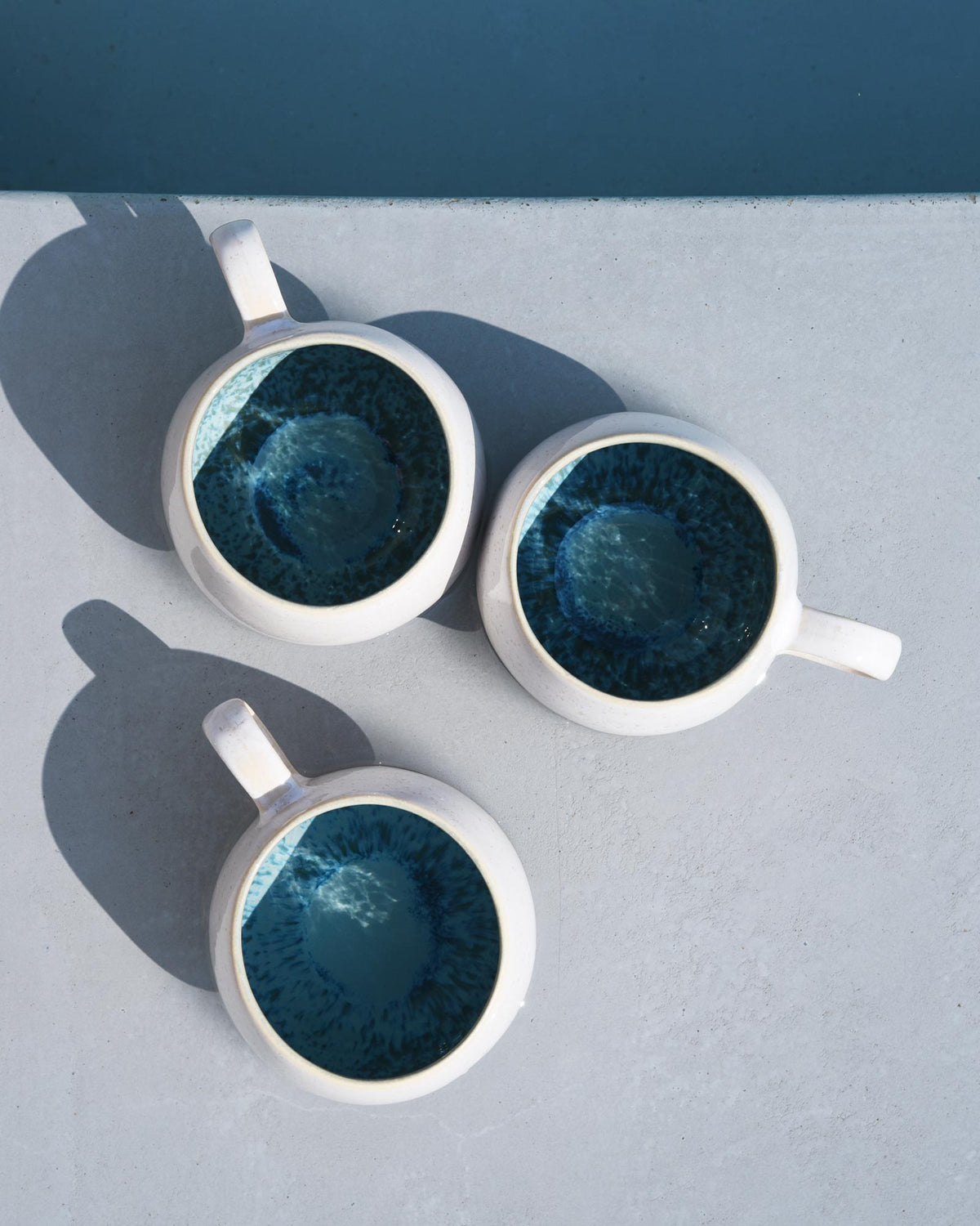Drei weiße Tassen mit dunkelblauer Innenseite stehen auf einem grauen Tisch, beleuchtet von hellem Sonnenlicht.