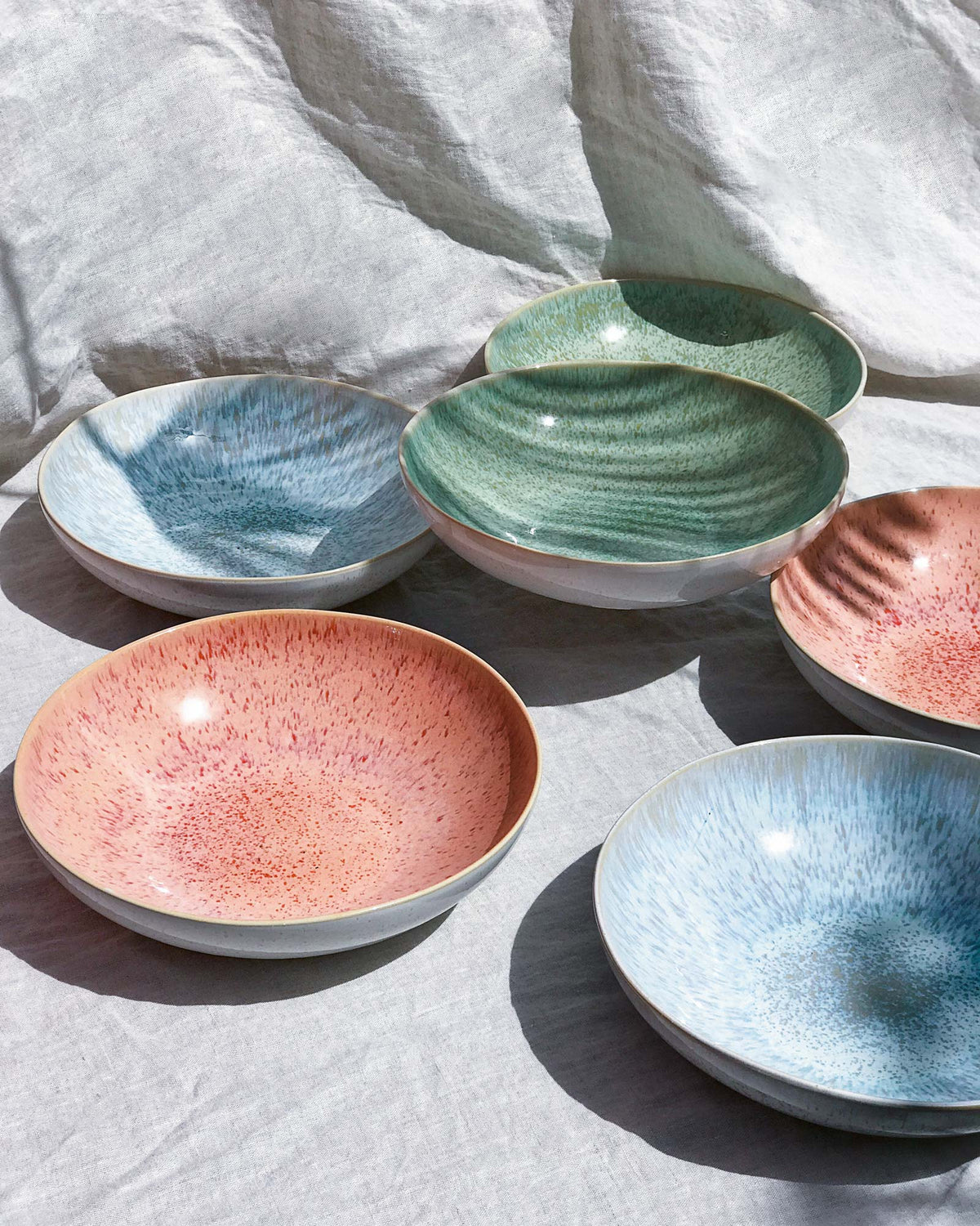 Sechs bunte Keramikschalen in Blau, Grün und Rosa stehen auf einem strukturierten weißen Hintergrund.