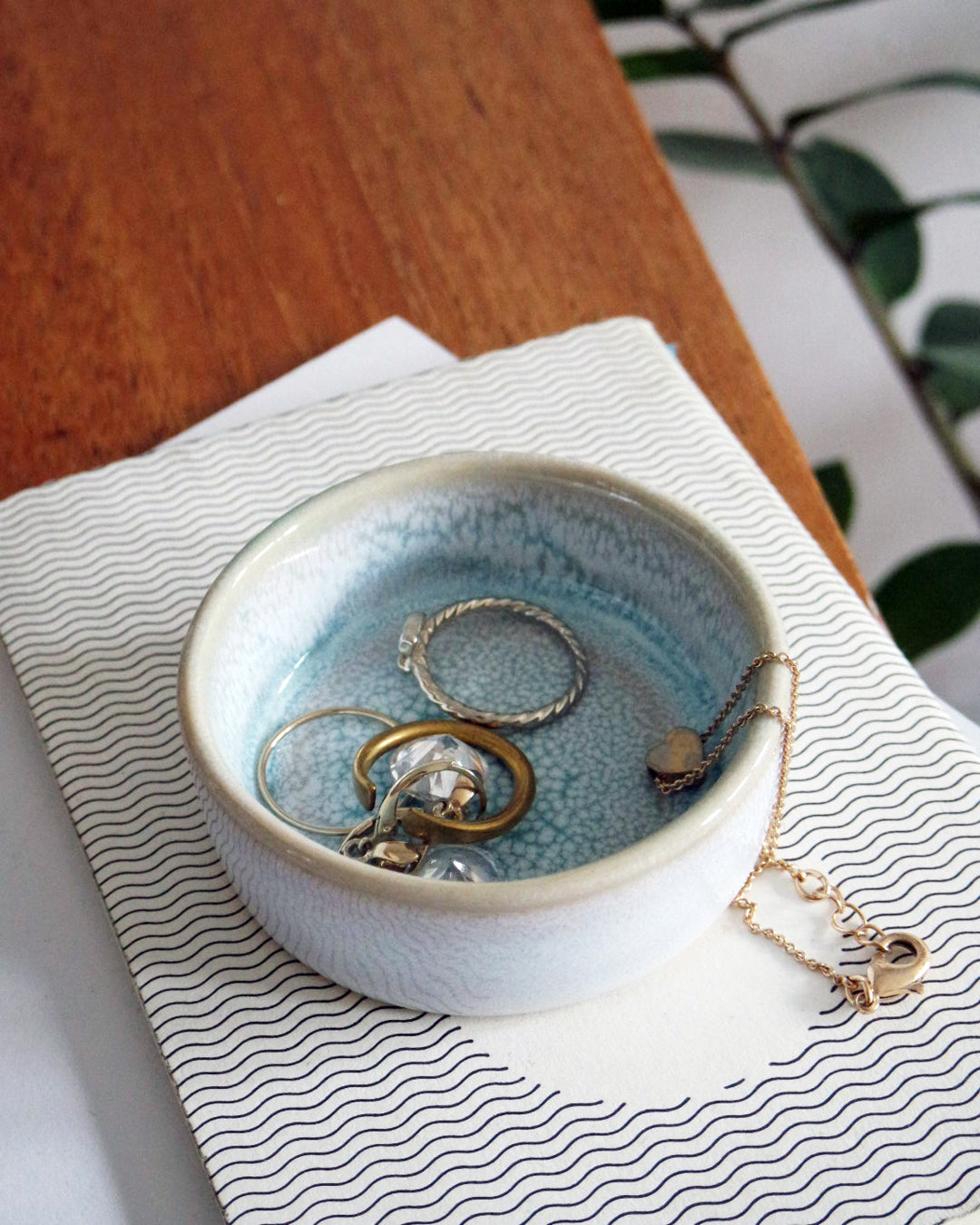 Eine kleine, blaue Keramikschale mit Schmuck auf einem Buch mit wellenförmigem Muster, auf einem Holztisch stehend.