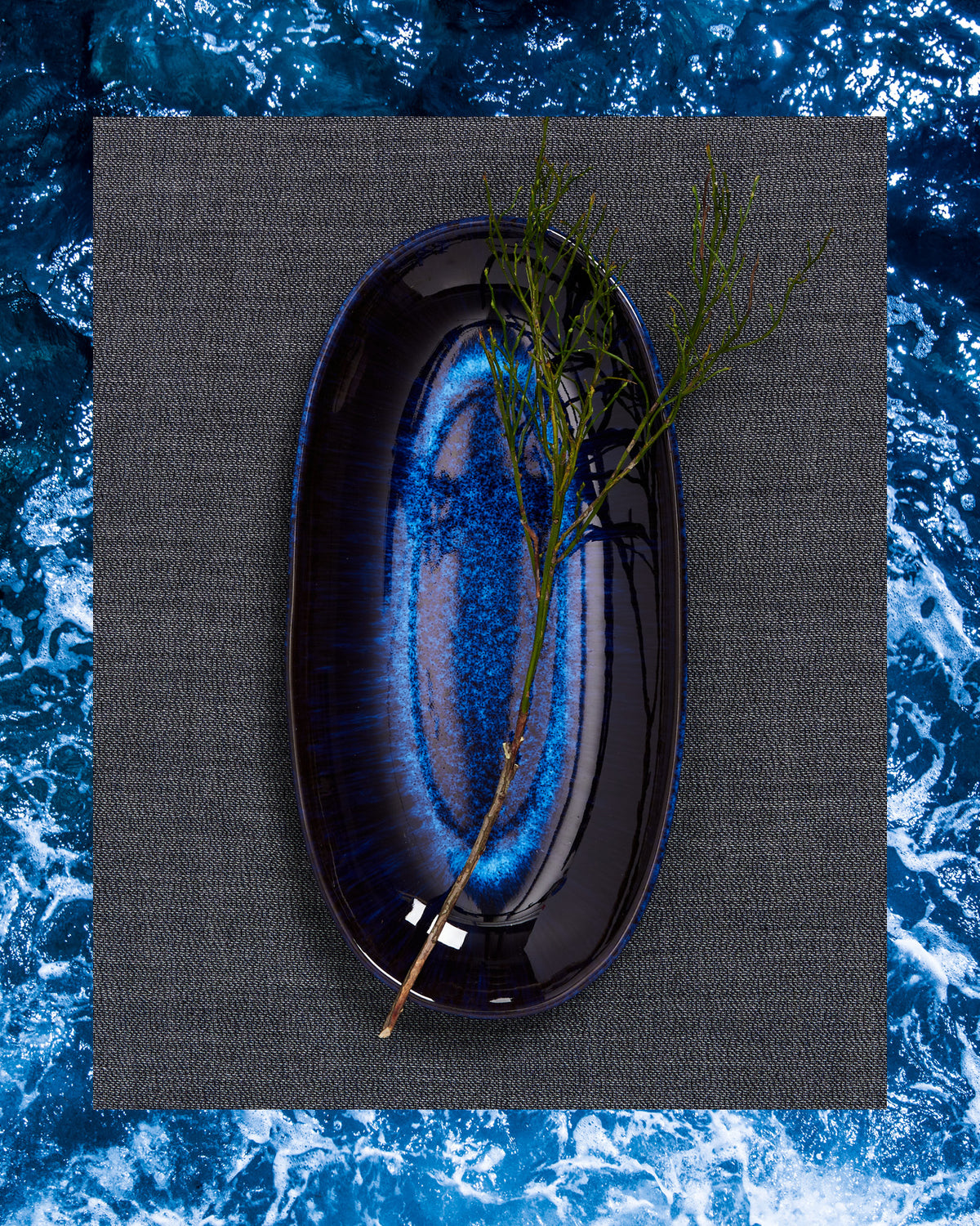 Blauer, ovaler Teller mit einem Zweig darauf, auf einem grauen Tuch und blauem Wasserhintergrund.