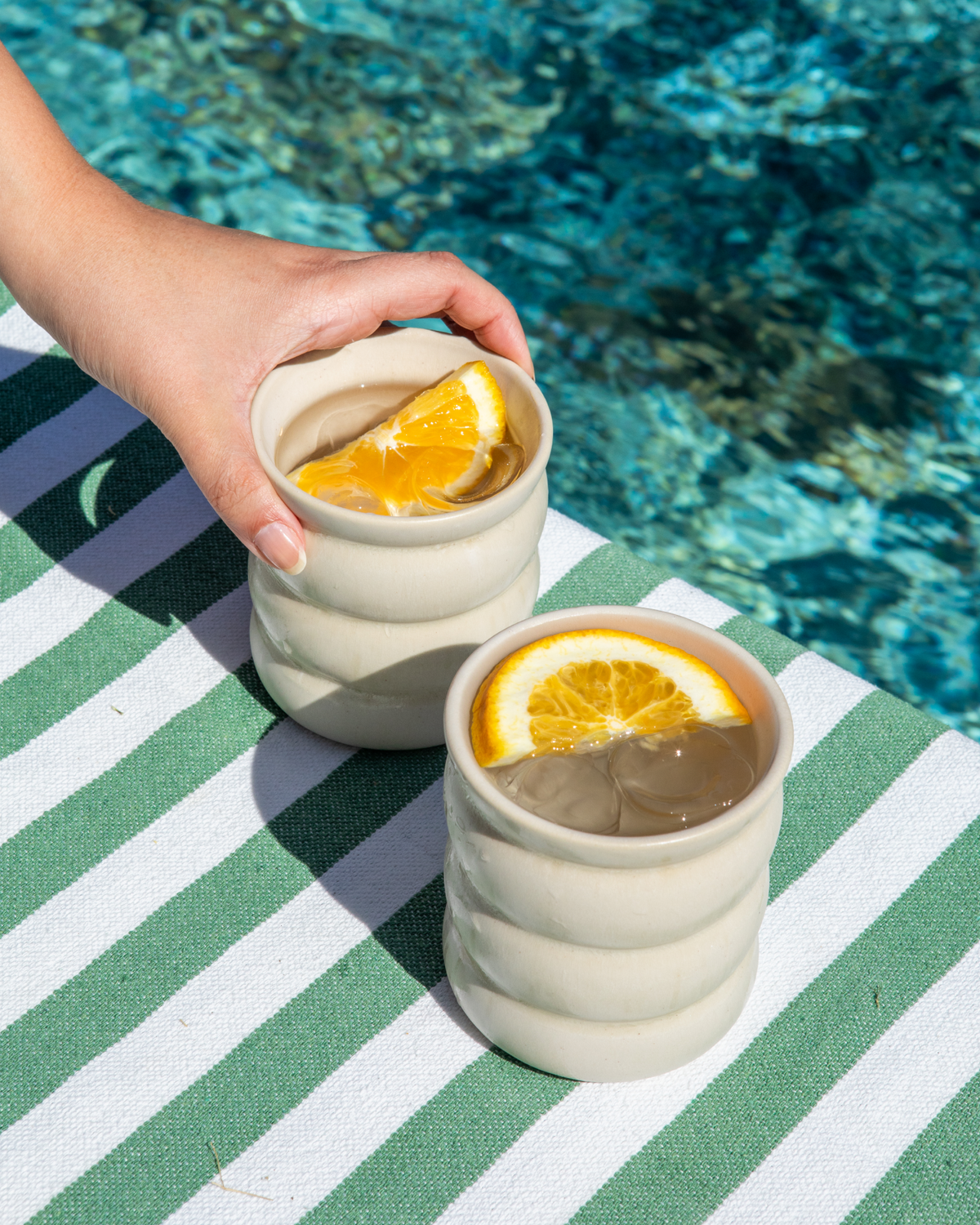 Eine Hand hebt einen Becher mit Eis und Orangenscheibe von einem grün-weiß gestreiften Handtuch am Poolrand.