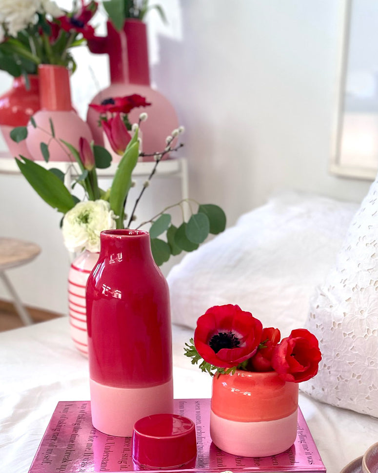 Rote und rosafarbene Vasen mit Blumen auf einem Tisch, im Hintergrund weitere Vasen und Pflanzen.